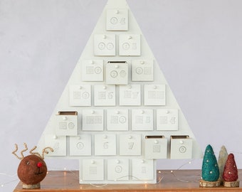 Pode incluir: Calendário do Advento de madeira branca em forma de árvore de Natal com 24 gavetas numeradas. A árvore está sobre uma mesa de madeira com um pequeno ornamento de rena e três pequenas árvores de Natal de feltro.