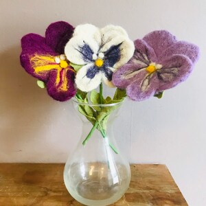 May include: Three felt pansies in a clear glass vase. The pansies are purple, white, and light purple. The pansies have yellow centers and dark purple markings.