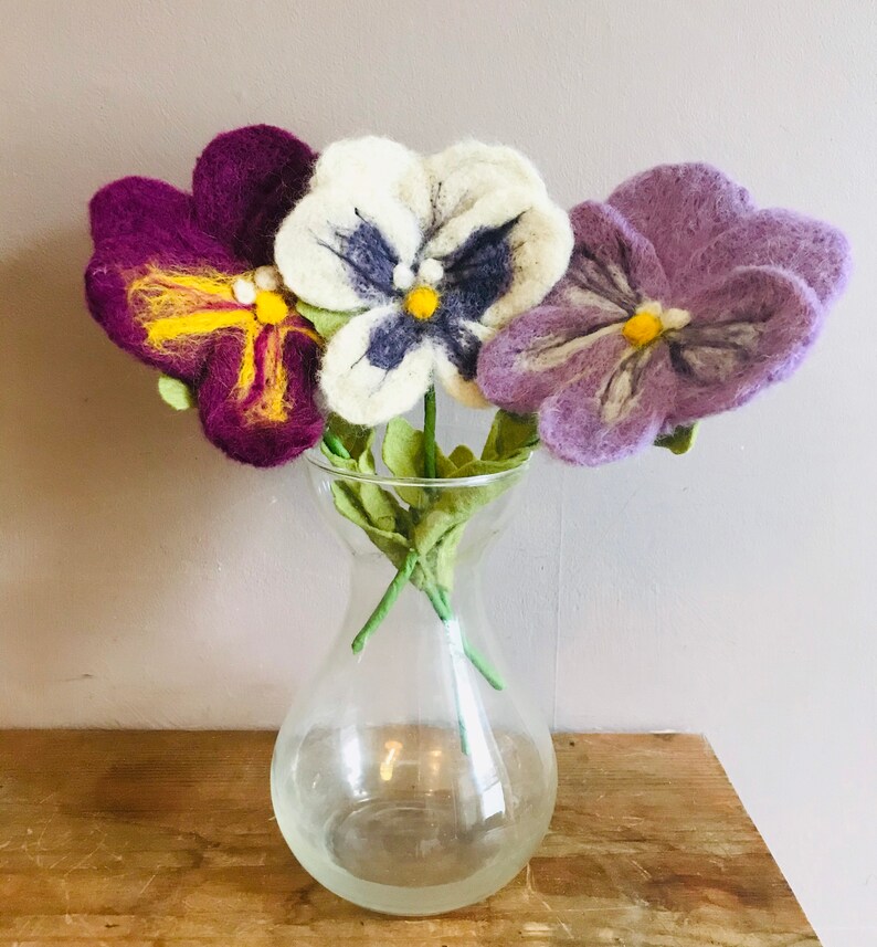 May include: Three felt pansies in a clear glass vase. The pansies are purple, white, and light purple. The pansies have yellow centers and dark purple markings.