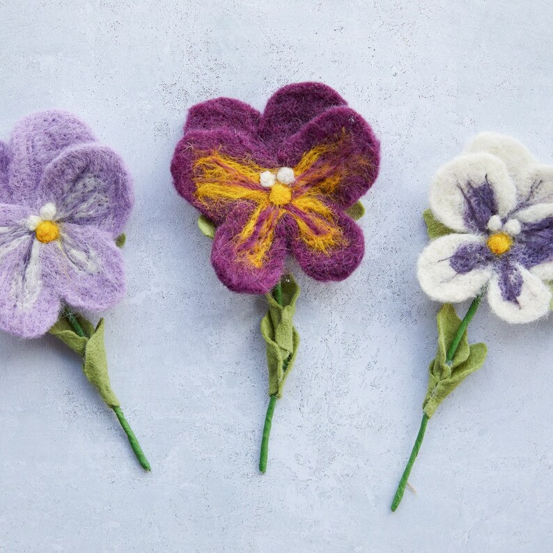 May include: Three felt flower pins in purple, white, and dark purple with yellow centers. The flowers are on green stems.