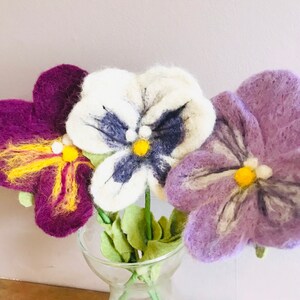 May include: Three felt pansies in a clear glass vase. The pansies are purple, white with blue and black accents, and light purple with white accents. The pansies have yellow centers.