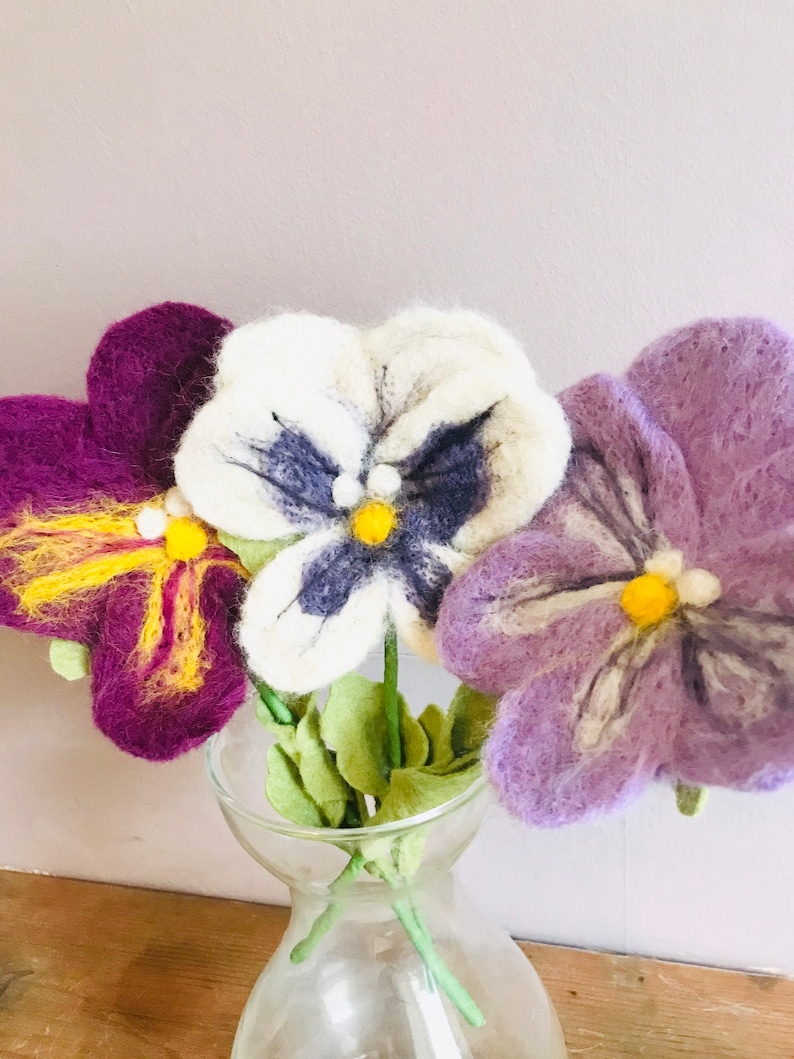 May include: Three felt pansies in a clear glass vase. The pansies are purple, white with blue and black accents, and light purple with white accents. The pansies have yellow centers.