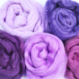 Puede incluir: Cinco ovillos de lana en tonos de morado, rosa y lavanda. La lana es suave y esponjosa, perfecta para tejer o hacer ganchillo.