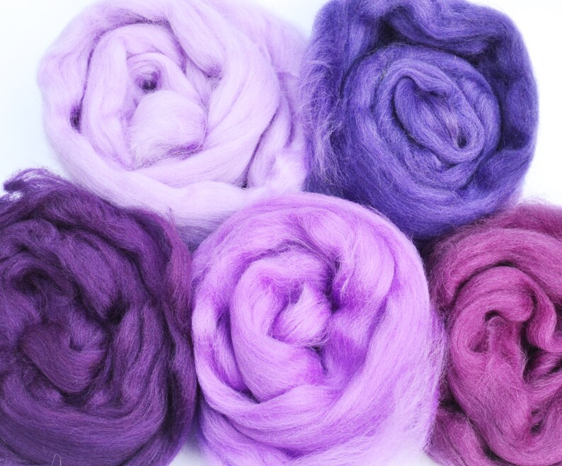 Puede incluir: Cinco ovillos de lana en tonos de morado, rosa y lavanda. La lana es suave y esponjosa, perfecta para tejer o hacer ganchillo.