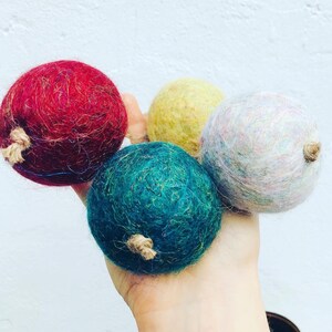 Pode incluir: Quatro bolas de lã feltradas em diferentes cores: vermelho, amarelo, branco e azul-petróleo. As bolas são seguradas na mão.