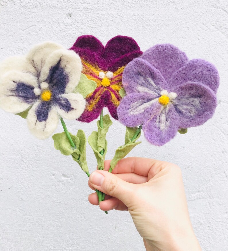 May include: Three felt pansies in white, purple, and burgundy with yellow centers. The flowers are attached to green stems.