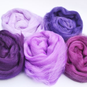 Puede incluir: Cinco ovillos de lana en tonos de morado, lavanda y rosa. La lana es suave y esponjosa, perfecta para tejer o hacer ganchillo.