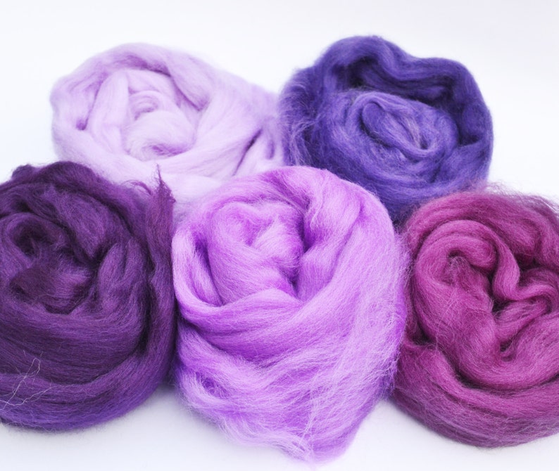 Puede incluir: Cinco ovillos de lana en tonos de morado, lavanda y rosa. La lana es suave y esponjosa, perfecta para tejer o hacer ganchillo.