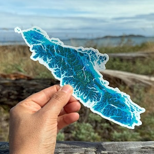 May include: A sticker in the shape of a landmass, featuring a detailed blue and green topographical design. The sticker is held against a backdrop of a blue sky and ocean, suggesting an outdoor theme.