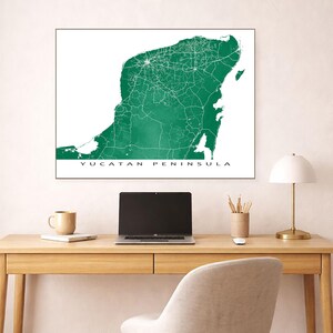 May include: A framed map of the Yucatan Peninsula in a vibrant green hue, showcasing the region's roads and cities in white. The text "YUCATAN PENINSULA" is printed in a clean, sans-serif font at the bottom. The map is displayed on a desk with a laptop, lamp, and other office supplies.