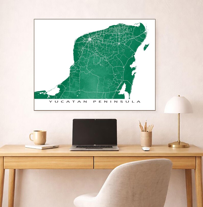 May include: A framed map of the Yucatan Peninsula in a vibrant green hue, showcasing the region's roads and cities in white. The text "YUCATAN PENINSULA" is printed in a clean, sans-serif font at the bottom. The map is displayed on a desk with a laptop, lamp, and other office supplies.