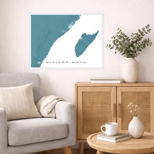 May include: A framed wall art print featuring a teal map of the Riviera Maya coastline. The print has white text that reads "RIVIERA MAYA". The artwork is displayed above a gray armchair and a wooden cabinet with a vase of greenery.