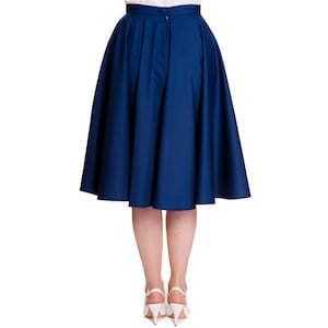 May include: A navy blue, knee-length skirt with a full, flared silhouette. The skirt features a high waist and a zipper closure at the back. The model is wearing white heels. The skirt is made of a solid, opaque fabric.