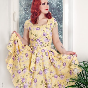 May include: A yellow dress featuring a square neckline, short sleeves, and a waist tie. The fabric is decorated with a repeating pattern of purple roses and green leaves. The skirt is flared and reaches knee length, showcasing a floral print.