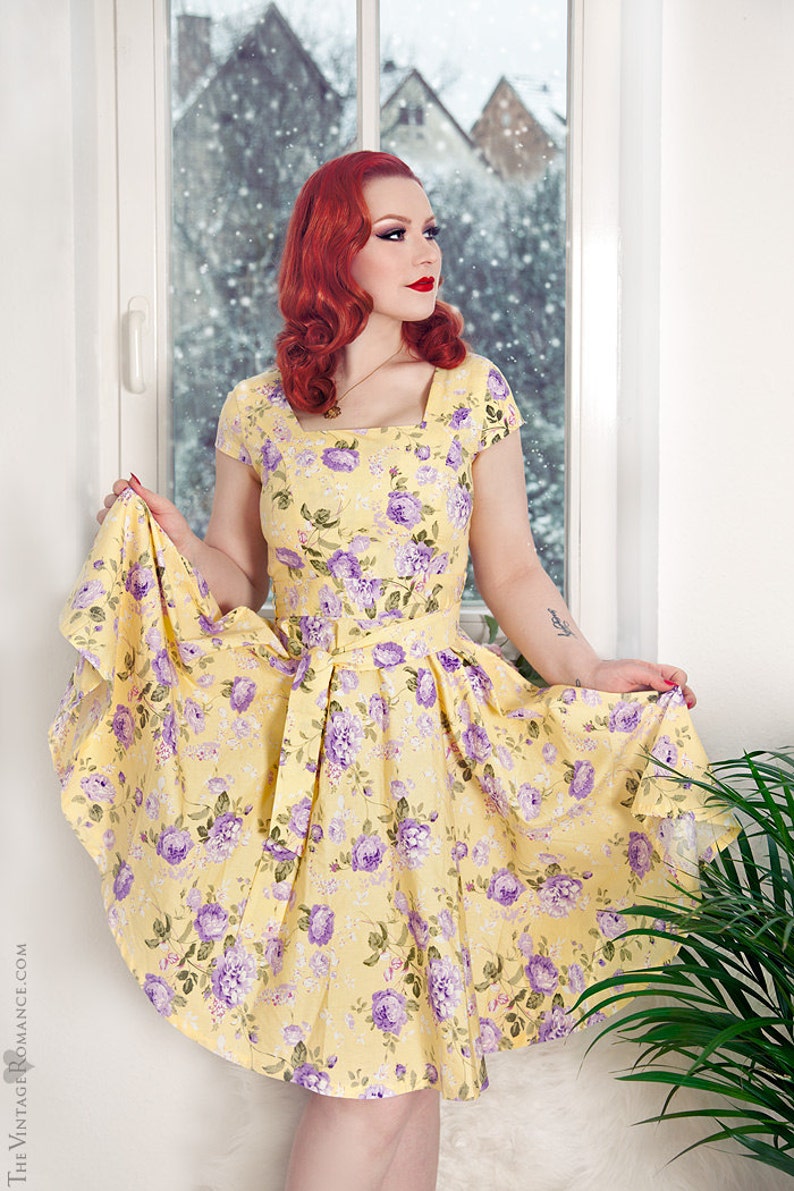 May include: A yellow dress featuring a square neckline, short sleeves, and a waist tie. The fabric is decorated with a repeating pattern of purple roses and green leaves. The skirt is flared and reaches knee length, showcasing a floral print.