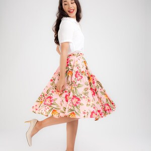 Puede incluir: Una mujer con una camisa blanca con cuello y una falda rosa con flores. La falda presenta un patr&oacute;n brillante de flores rojas, rosas y verdes. Lleva tacones beige. El atuendo es adecuado para una ocasi&oacute;n informal o semiformal.