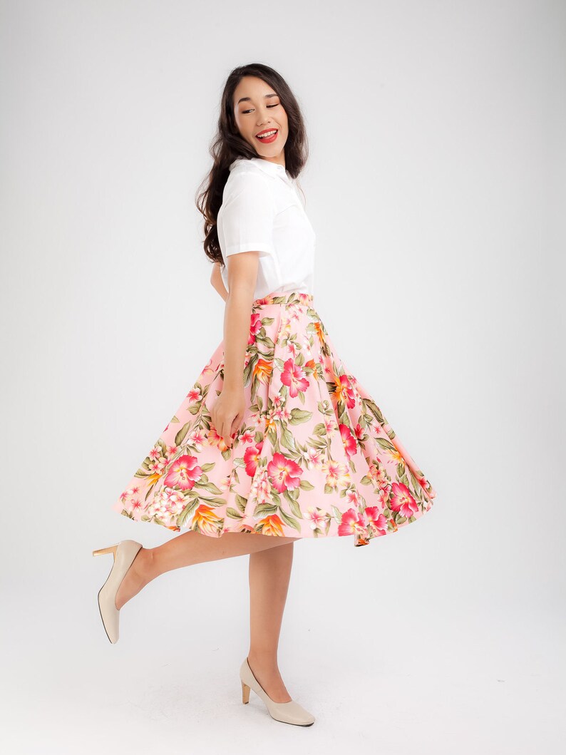 Puede incluir: Una mujer con una camisa blanca con cuello y una falda rosa con flores. La falda presenta un patr&oacute;n brillante de flores rojas, rosas y verdes. Lleva tacones beige. El atuendo es adecuado para una ocasi&oacute;n informal o semiformal.