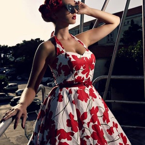 K&ouml;nnte beinhalten: Ein wei&szlig;es Neckholder-Kleid mit einem kr&auml;ftigen roten Blumenmuster, einem herzf&ouml;rmigen Ausschnitt und einem d&uuml;nnen roten G&uuml;rtel mit herzf&ouml;rmiger Schnalle. Das Kleid hat einen weit ausgestellten Rock und wird mit einer dunklen Cat-Eye-Sonnenbrille kombiniert.