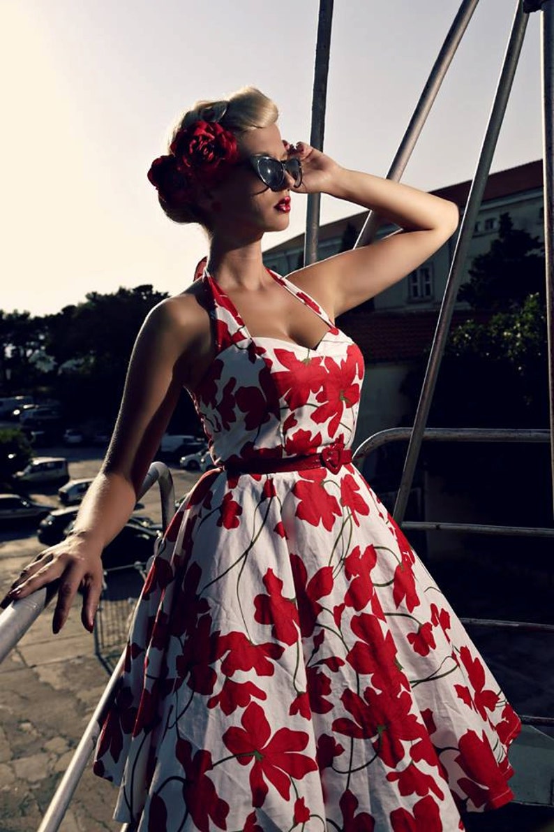 K&ouml;nnte beinhalten: Ein wei&szlig;es Neckholder-Kleid mit einem kr&auml;ftigen roten Blumenmuster, einem herzf&ouml;rmigen Ausschnitt und einem d&uuml;nnen roten G&uuml;rtel mit herzf&ouml;rmiger Schnalle. Das Kleid hat einen weit ausgestellten Rock und wird mit einer dunklen Cat-Eye-Sonnenbrille kombiniert.