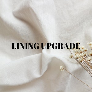 May include: A close-up of white fabric with the words "LINING UPGRADE" in black. A small bundle of dried white flowers is in the lower right corner. The fabric is draped and has soft folds.
