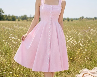 Puede incluir: Un vestido sin mangas de color rosa claro con un sutil estampado de cuadros vichy blancos, escote cuadrado y falda con vuelo. El corpiño se cierra con una hilera vertical de botones blancos en el centro y el vestido incluye bolsillos laterales.