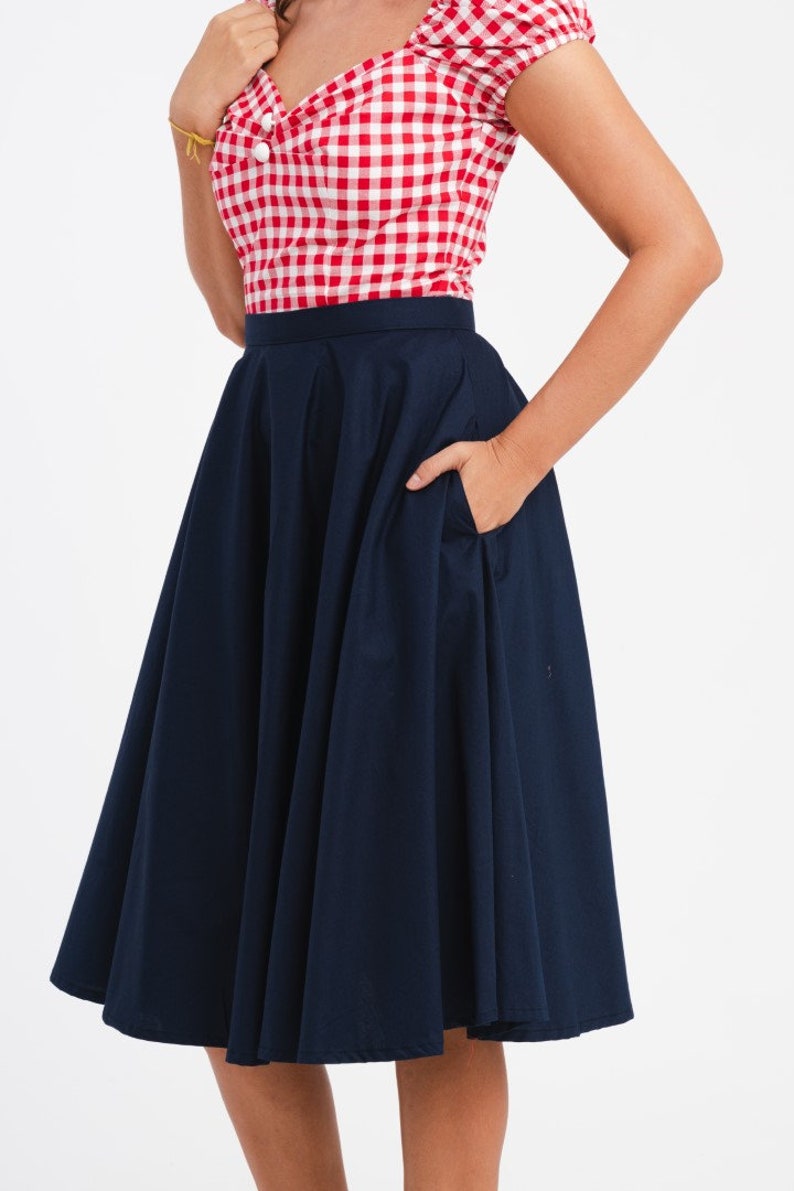 May include: A red and white checkered top with a navy blue skirt. The skirt is a full, flared style with a pocket. The top has a square neckline and white buttons. The outfit is a classic, retro style.