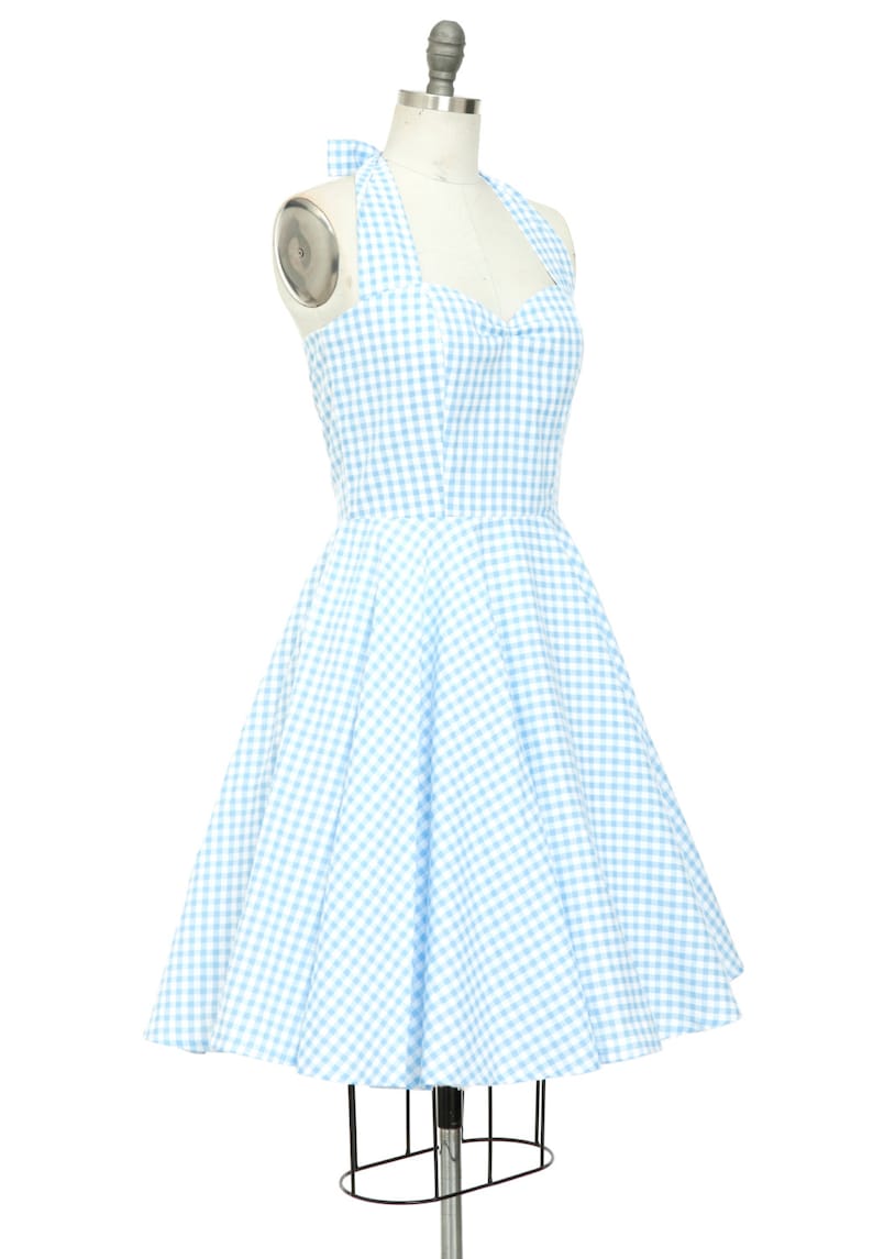 K&ouml;nnte beinhalten: Hellblau-wei&szlig;es Gingham-Halterkleid auf einer Schaufensterpuppe. Das Kleid hat ein figurbetontes Oberteil, einen ausgestellten Rock und eine Schleife am Nacken. Das Kleid ist ein klassischer, vintage-inspirierter Stil.