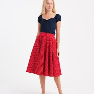 May include: A woman wearing a navy blue top with short sleeves and a sweetheart neckline. She is also wearing a red, knee-length skirt with white polka dots. The outfit is paired with white slip-on shoes. The background is white.
