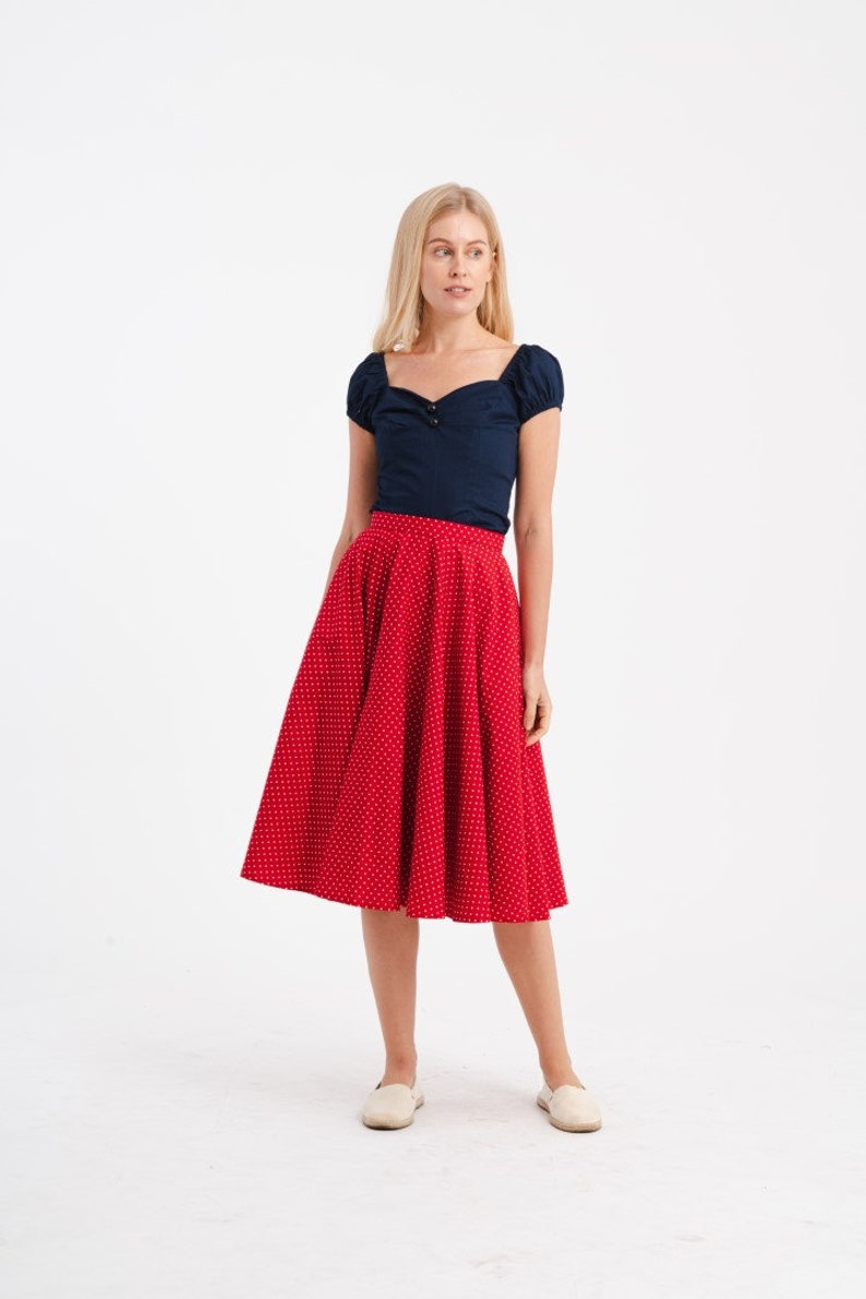 May include: A woman wearing a navy blue top with short sleeves and a sweetheart neckline. She is also wearing a red, knee-length skirt with white polka dots. The outfit is paired with white slip-on shoes. The background is white.