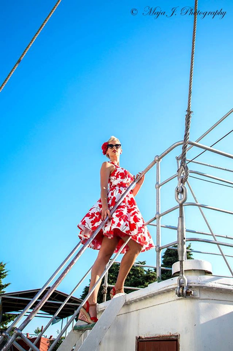 K&ouml;nnte beinhalten: Ein &auml;rmelloses wei&szlig;es Kleid mit rotem Blumenmuster, weitem Rock und eng anliegendem Oberteil. Das Outfit umfasst rote Keilsandaletten, eine dunkle Sonnenbrille und ein gro&szlig;es rotes Blumen-Haaraccessoire, getragen auf einem Schiffsdeck.
