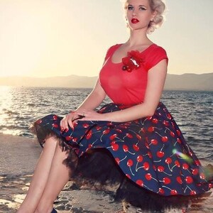 Puede incluir: Una mujer con top rojo y falda azul marino con estampado de cerezas, ribeteada con encaje negro. Lleva bailarinas azul marino. El fondo muestra el mar y una puesta de sol. Estilo retro.