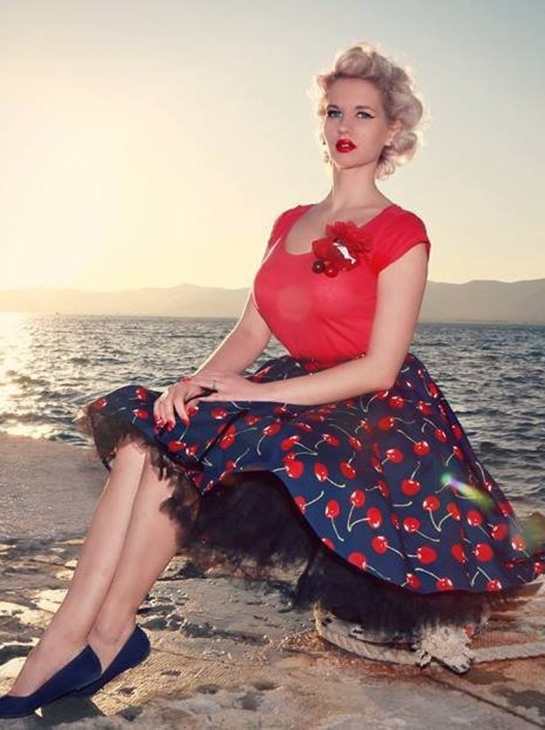 Puede incluir: Una mujer con top rojo y falda azul marino con estampado de cerezas, ribeteada con encaje negro. Lleva bailarinas azul marino. El fondo muestra el mar y una puesta de sol. Estilo retro.