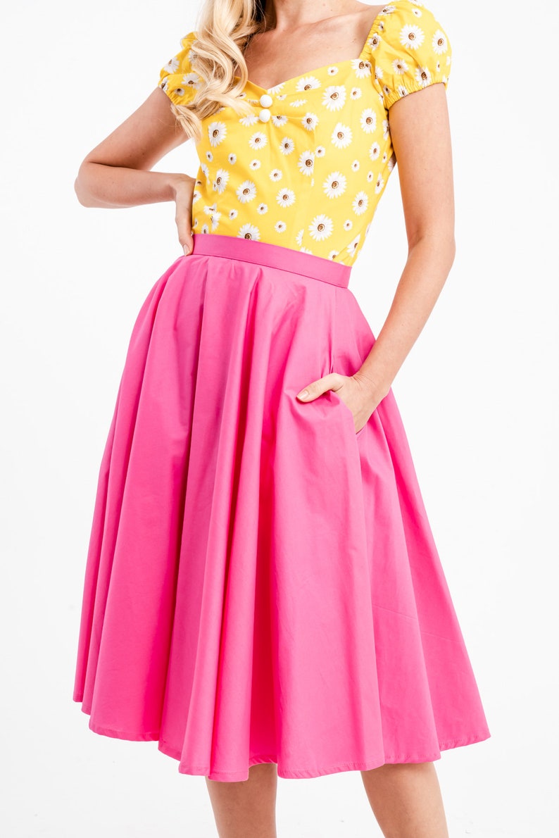 Puede incluir: Una blusa amarilla brillante con estampado de margaritas combinada con una falda rosa vibrante. La falda tiene una cintura ancha y una forma amplia y fluida. El conjunto es alegre y femenino.