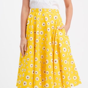 May include: A yellow skirt with a white daisy pattern. The skirt has a flared design and pockets. The model is wearing a white top with short sleeves. The skirt is a knee-length style.