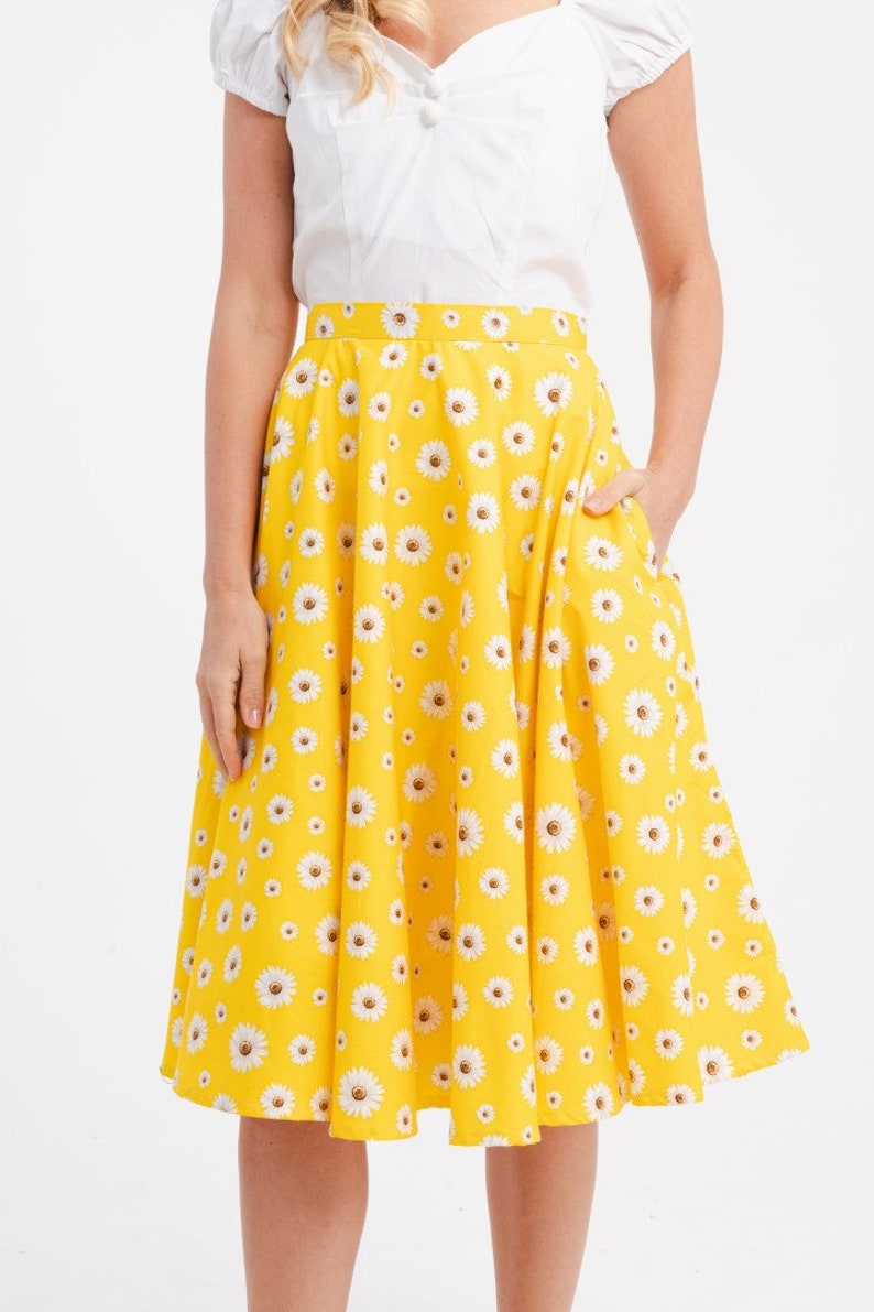 May include: A yellow skirt with a white daisy pattern. The skirt has a flared design and pockets. The model is wearing a white top with short sleeves. The skirt is a knee-length style.