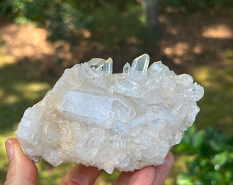 May include: A cluster of clear quartz crystals with a rough, natural texture. The crystals are arranged in a random pattern, with some pointing upwards and others pointing downwards. The crystals are a light white color and have a slight sparkle.