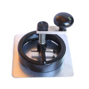 May include: A metal circle cutter with a black handle and knob, mounted on a silver square base. The cutter has a spring and a small black knob on top. The main knob is black and round.