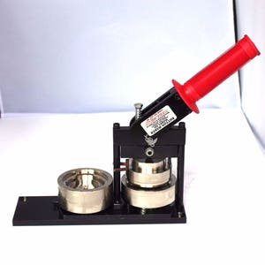 May include: A black button maker with a red handle. The machine has a metal base and a metal press. The machine is labeled "Button Maker Model 1000".