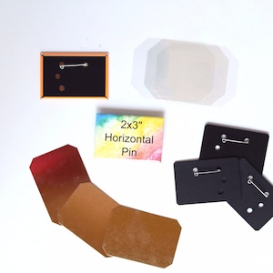 May include: An assortment of pin-making supplies, including black pin backs, a rectangular pin with the text "2x3" Horizontal Pin", and a folded brown card. The pin backs have a safety pin style clasp.