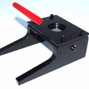 May include: Black metal button badge maker with a red lever. The machine has a circular die for making badges.