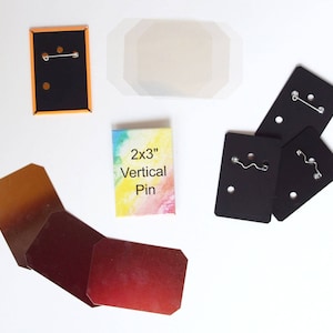 May include: An assortment of pin-back buttons and backing cards. The buttons include a black button with an orange border, and several black backing cards with pin fasteners. A colorful card reads "2x3" Vertical Pin."