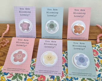 May include: Six colorful flower-shaped pins with the text "You Are Blooming Lovely!" on pink, purple, and blue cards. The cards have the text "@shaniceenglandtextiles" and "www.shaniceenglandart.etsy.com" printed on them.