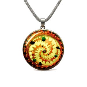 Pode incluir: Colar com pingente redondo e corrente prateada. O pingente apresenta um design circular com fundo amarelo-esverdeado, detalhes em laranja e vermelho, e enfeites verdes e pretos. O pingente tem aproximadamente 3,8 cm de diâmetro.