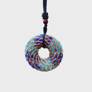 Op de afbeelding: Een cirkelvormige hanger met een spiraalvormig ontwerp, met blauwe en turquoise stenen ingekapseld in koperdraad. De hanger hangt aan een zwart koord met een houten kraal accent. Het ontwerp heeft een natuurlijke, handgemaakte uitstraling.