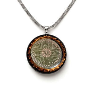 May include: A round pendant necklace with a silver chain. The pendant has a dark brown and gold rim, surrounding a green and gold circular design with intricate patterns. The necklace is displayed on a white background.