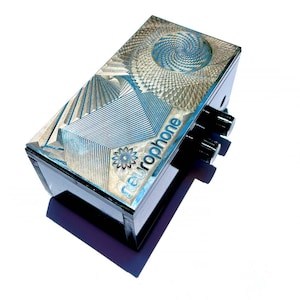 Puede incluir: Una caja negra con un diseño geométrico plateado y azul y la palabra "neurophone" impresa en la parte superior. La caja tiene dos botones en el lateral.