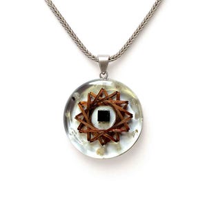 Könnte beinhalten: Ein klarer Harzanhänger mit einem kupferfarbenen Sternmuster und einem schwarzen quadratischen Mittelteil. Der Anhänger hängt an einer silbernen Kette. Das Design ist in klarem, kreisförmigem Harz eingeschlossen.