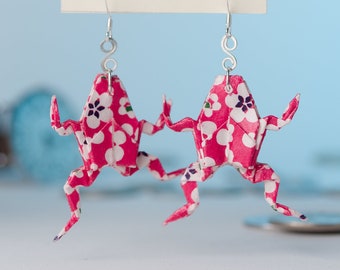 May include: A pair of origami frog earrings with a pink and white floral pattern. The earrings are made of paper and have silver hooks.