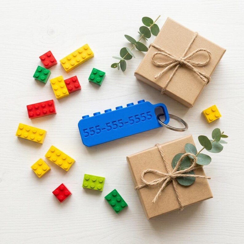 May include: A blue rectangular plastic keychain with the embossed text 555-555-5555 and a metal ring, surrounded by scattered red, yellow, and green plastic building blocks, two brown gift boxes tied with twine, and small green eucalyptus sprigs.