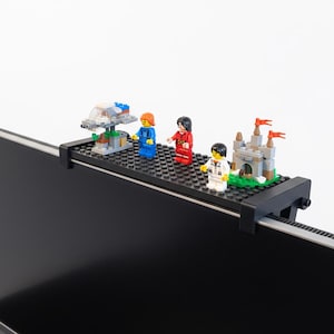 Puede incluir: Una estantería negra para monitor con una superficie negra con tachuelas sostiene varias estructuras y minifiguras de Lego. Las creaciones de Lego incluyen una nave espacial, un castillo y tres minifiguras con trajes azules, rojos y blancos.